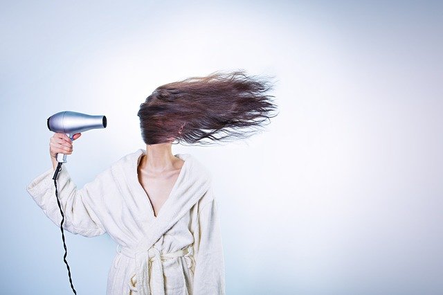 Rodzaje włosów: suche. Powinno unikać się suszenia ich suszarką. Na zdjęciu kobieta w szlafroku suszy włosy. 